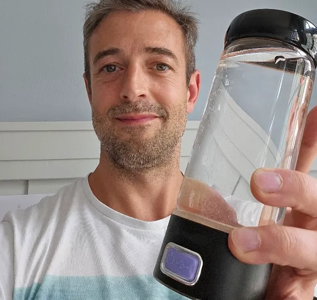 Marc Dietschi zeigt einen Wasser-Ionisator in die Kamera, sportlich gekleidet, mit einem hellen Hintergrund.