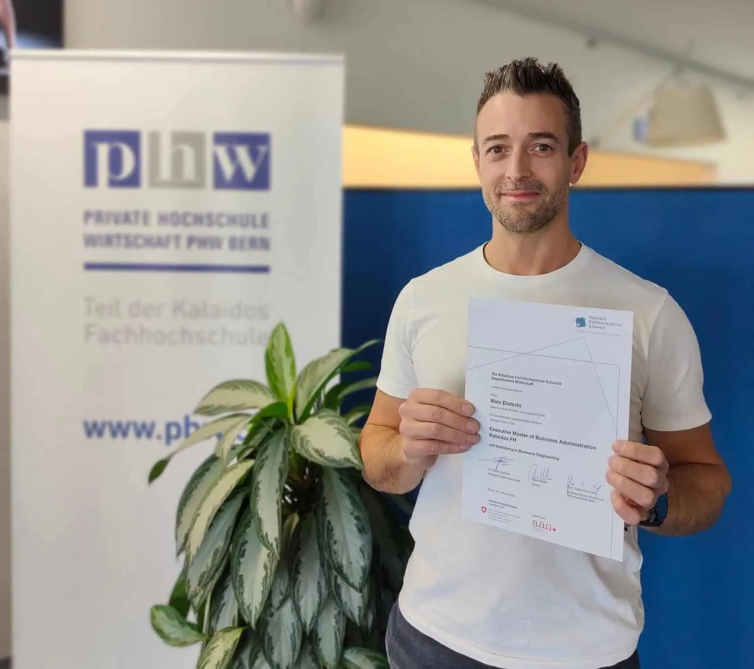 Marc Dietschi vor dem Logo der PHW Bern mit dem MBA-Diplom in seinen Händen.