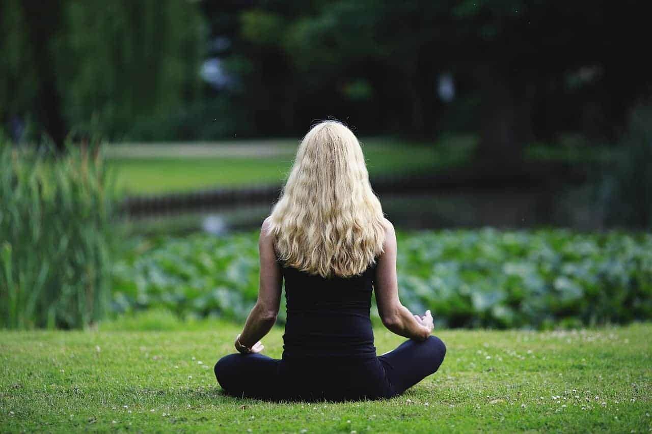 Frau mit blonden Haaren meditiert im Schneidersitz auf einer grünen Wiese, unscharfer Hintergrund mit Pflanzen.