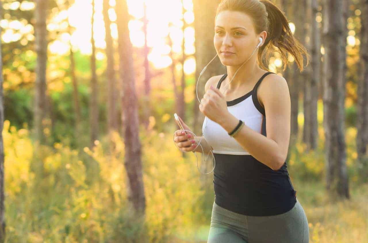 Ein sonnendurchfluteter Waldweg, auf dem eine Person im unscharfen Hintergrund joggt, strahlt Gesundheit und Aktivität aus.