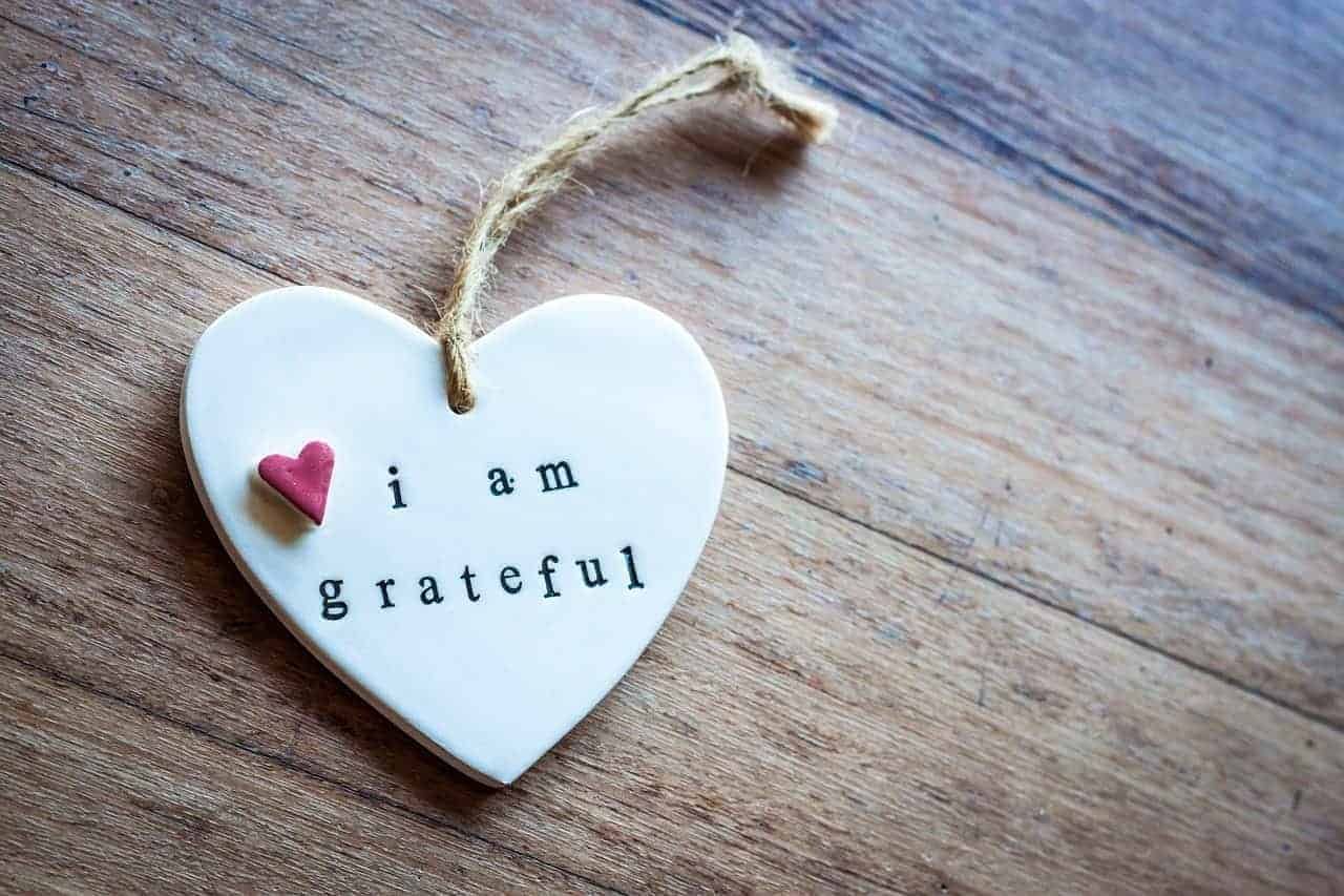 Herzförmiger Anhänger mit der Aufschrift "I am grateful" auf holzerner Oberfläche, mit kleinem rosa Herz oben links.