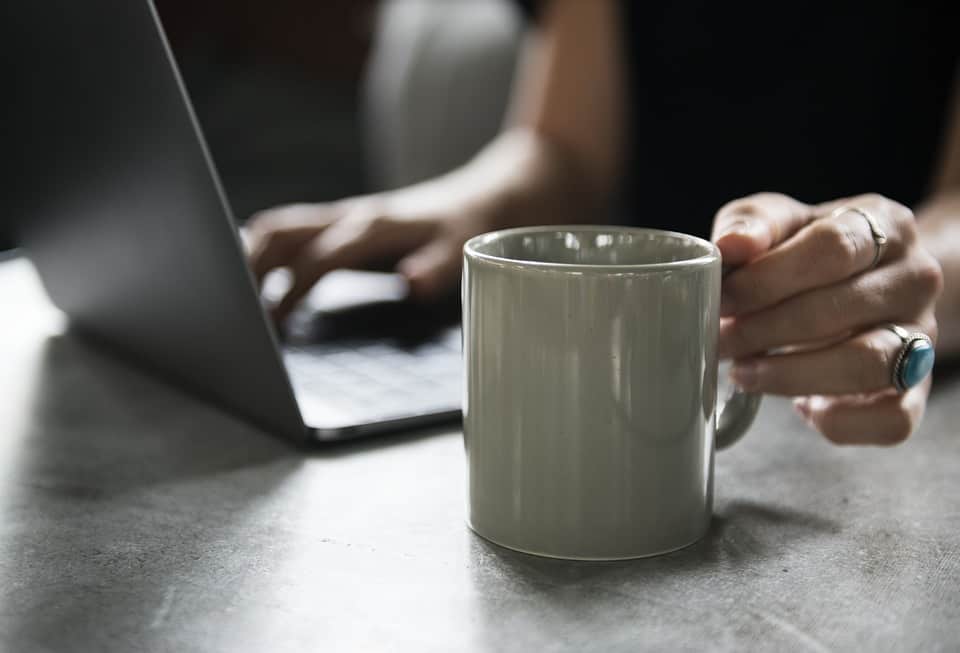 Eine graue Tasse mit Bulletproof Coffee und ein Notebook im Hintergrund. Eine Frau hat den Kaffee nach dem Rezept aus dem Internet hergestellt.