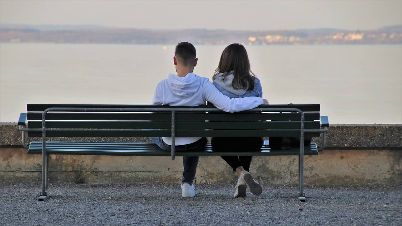 Ein Junge und ein Mädchen sitzen nebeneinander auf einer Bank und schauen entspannt dem Sonnenuntergang entgegen, da sie sich vertrauen.