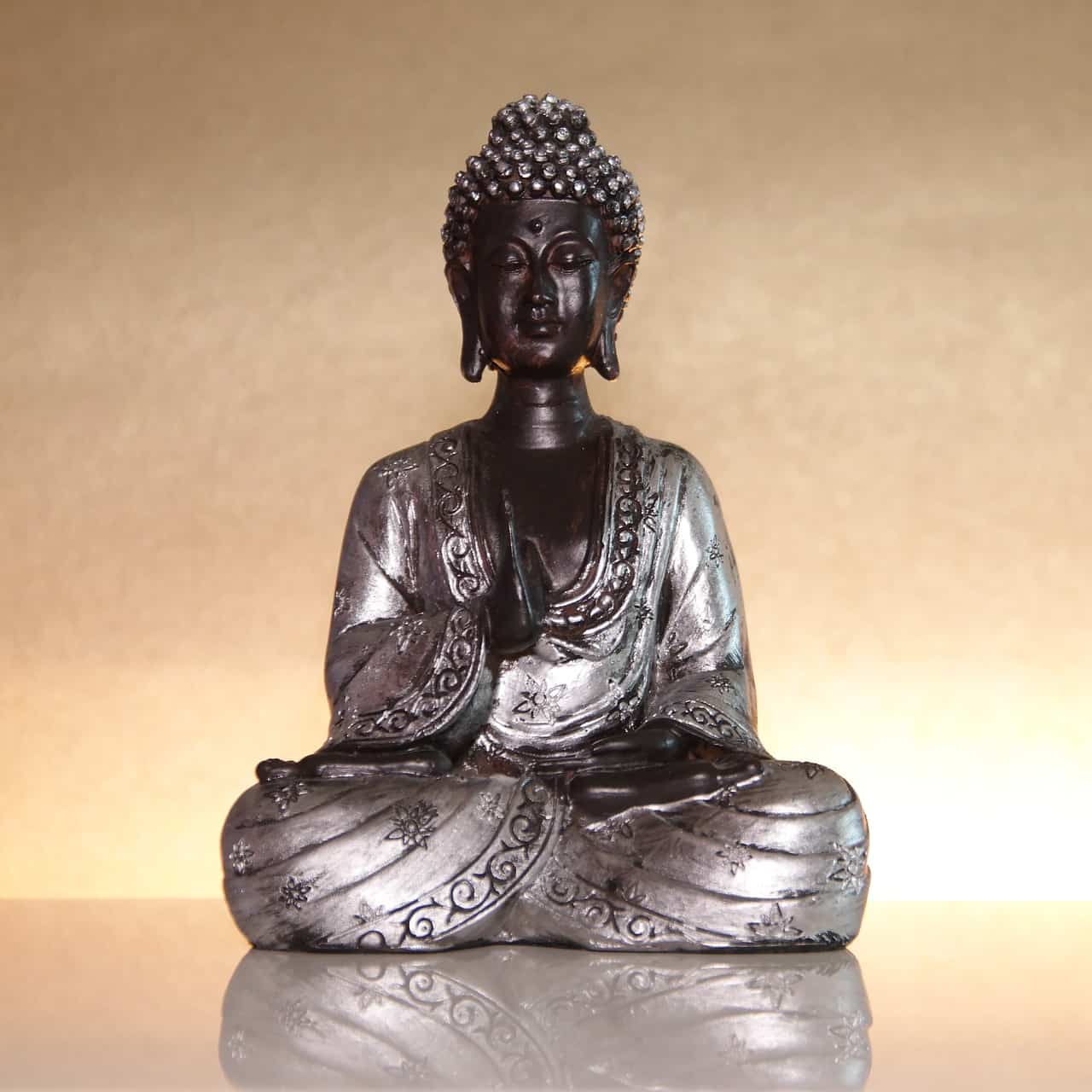 Silberne Buddha-Statue im Lotossitz sitzt auf einer spiegelten Oberfläche und goldener Rückwand.